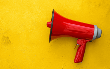 Ein roter Megafon liegt horizontal auf einem leuchtend gelben, leicht strukturierten Hintergrund. Der Megafon ist glänzend rot mit grauen Akzenten und einem Pistolengriff. Der starke Farbkontrast zwischen dem Rot und Gelb sowie die minimalistische Komposition erzeugen eine dynamische, aufmerksamkeitsstarke Wirkung.