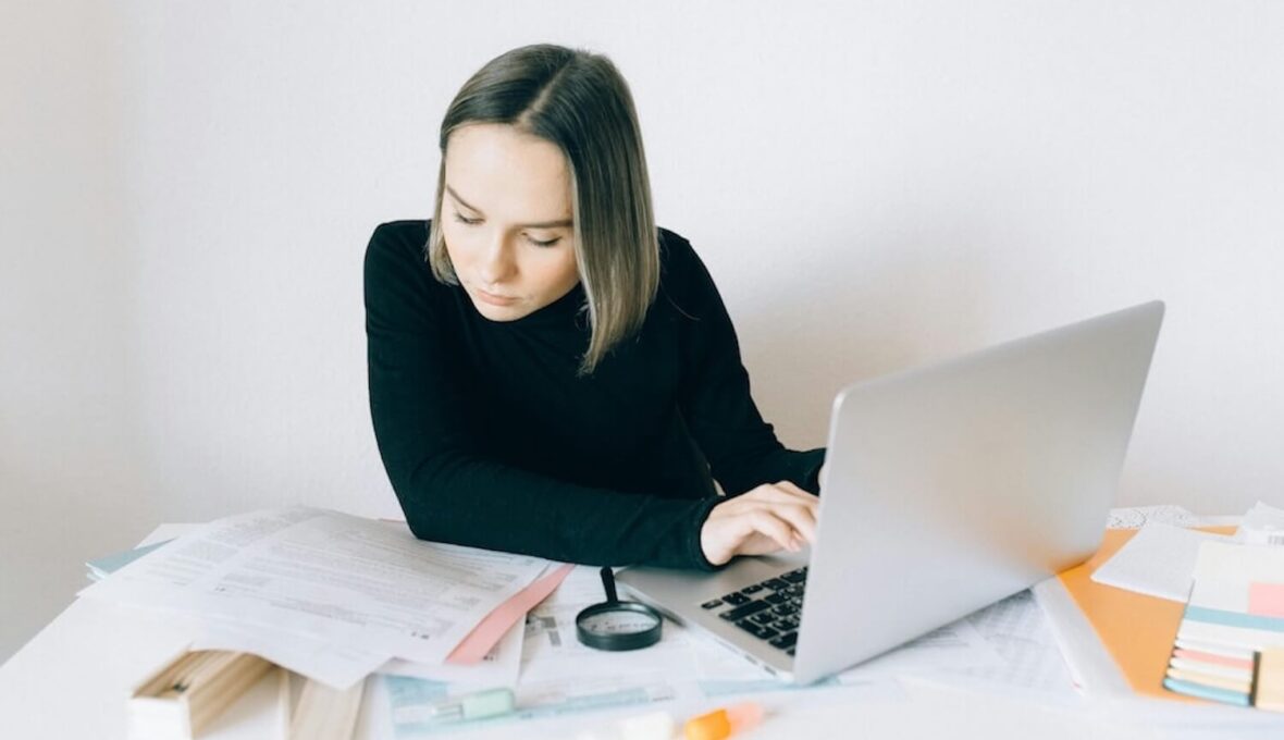 Frau sitzt an einem Schreibtisch, arbeitet konzentriert an einem Laptop und ist von Dokumenten umgeben. Sie trägt einen schwarzen Rollkragenpullover. Im Hintergrund ist eine helle, minimalistische Wand.