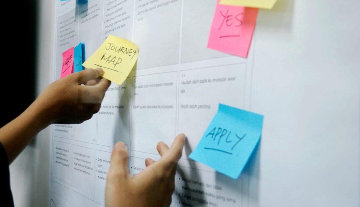 Zwei Hände platzieren bunte Haftnotizen auf einer weißen Planungstafel oder einem Whiteboard. Auf den sichtbaren Notizzetteln stehen Worte wie "JOURNEY MAP" (gelb) und "APPLY" (blau). Die Tafel enthält gedruckten Text und Linien im Hintergrund, die auf eine strukturierte Projektplanung oder einen Workflow hinweisen. Die Notizzettel sind in den Farben Gelb, Rosa und Blau.